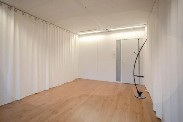 Lichte, lege kamer met houten vloer, witte gordijnen en een moderne kapstok bij een gesloten deur.